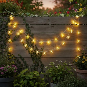May include: A string of flower-shaped fairy lights illuminates a wooden fence. The warm yellow lights are strung across the fence, creating a soft glow. Green plants and flowers surround the fence, adding a natural touch to the scene.
