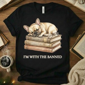 Puede incluir: Camiseta negra con un bulldog francés durmiendo sobre una pila de libros con gafas. Los libros se titulan "1984", "The Catcher in the Rye" y "Fahrenheit 451". El texto "I'M WITH THE BANNED" está impreso debajo.