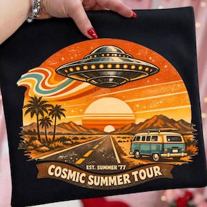 May include: Black t-shirt with a retro graphic of a UFO, a road, and a vintage van under a sunset. The text reads "Cosmic Summer Tour" and "Est. Summer '77". Palm trees and mountains are also featured.