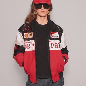May include: A red Ferrari baseball cap and a racing jacket with red, white, and black panels. The jacket features the Ferrari logo and sponsor patches. The person is wearing a black t-shirt and sunglasses.