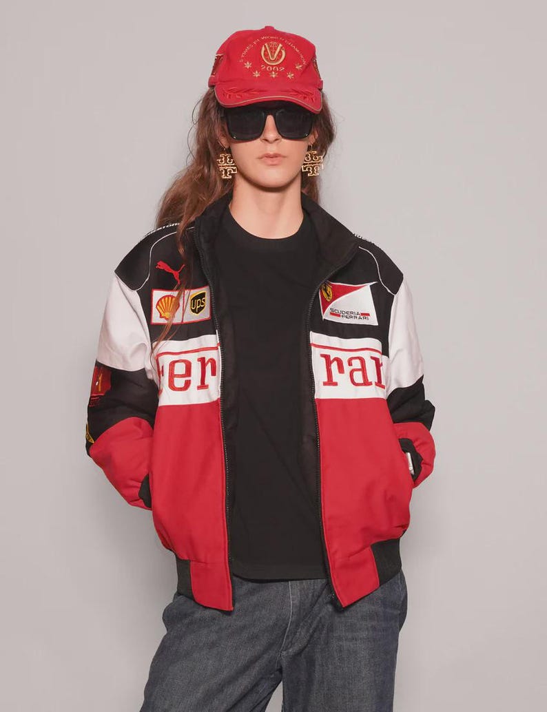 May include: A red Ferrari baseball cap and a racing jacket with red, white, and black panels. The jacket features the Ferrari logo and sponsor patches. The person is wearing a black t-shirt and sunglasses.