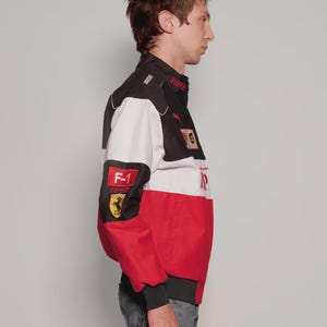 May include: A black, white, and red racing jacket featuring the Ferrari logo and F-1 patch. The jacket has a stand-up collar, ribbed cuffs, and a bomber-style fit. The jacket is made of a durable material.