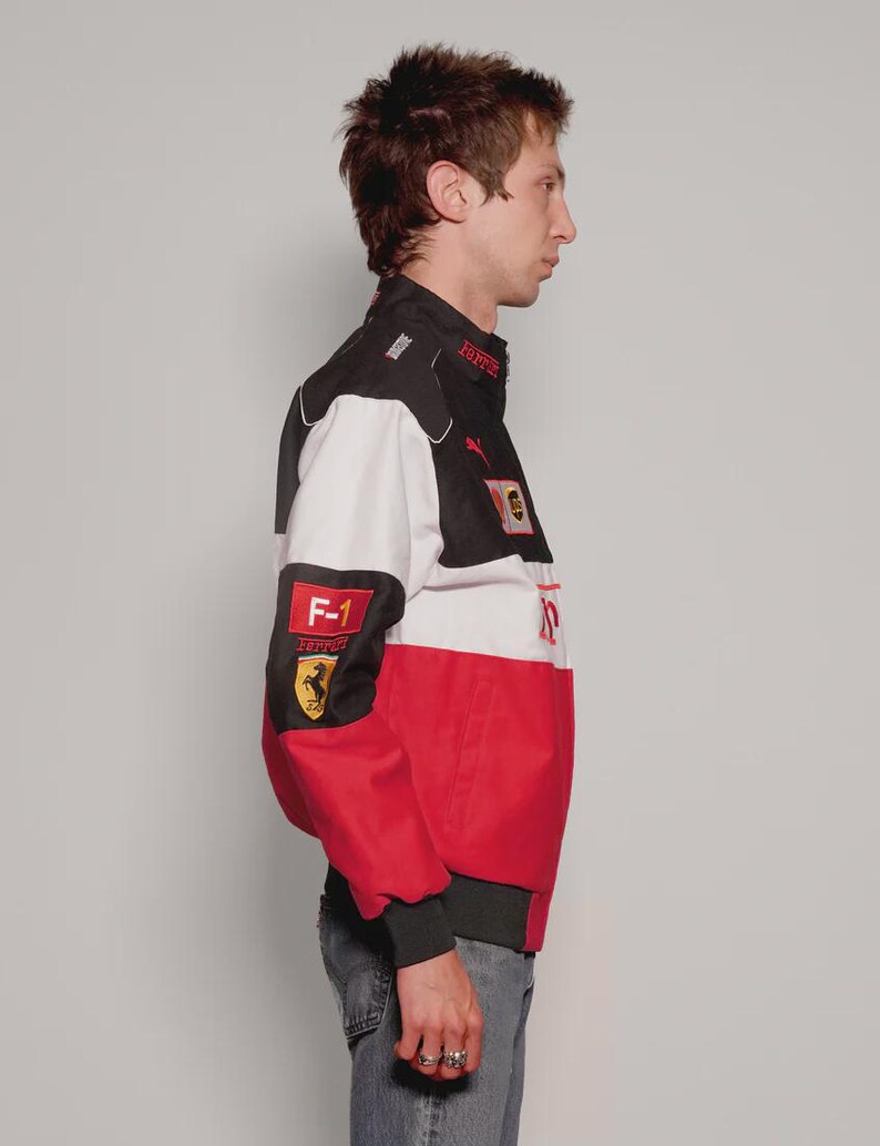 May include: A black, white, and red racing jacket featuring the Ferrari logo and F-1 patch. The jacket has a stand-up collar, ribbed cuffs, and a bomber-style fit. The jacket is made of a durable material.