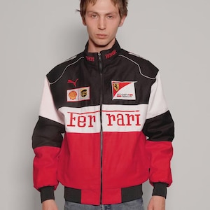 May include: A Ferrari racing jacket in black, white, and red, with a zip closure. The jacket displays the Ferrari logo, sponsor branding, and the word "Ferrari" in red across the chest. It has a ribbed collar and cuffs.