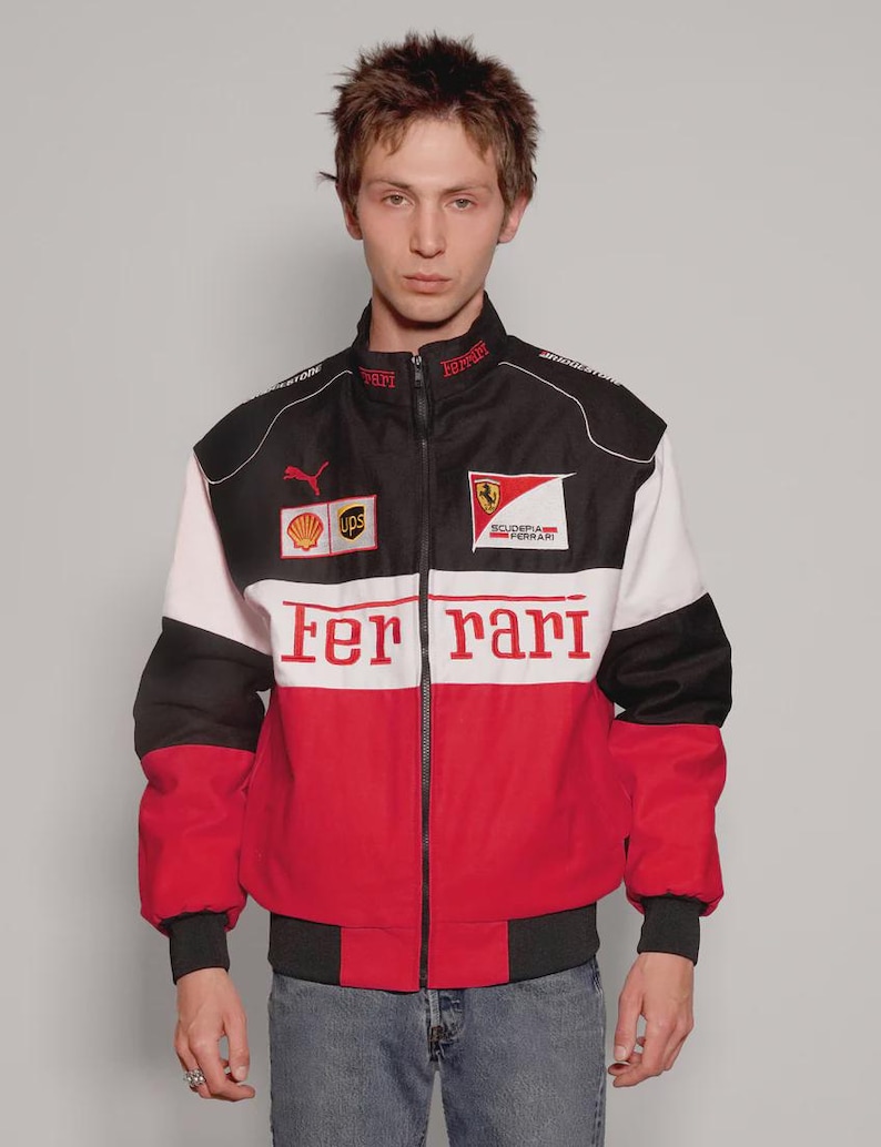 May include: A Ferrari racing jacket in black, white, and red, with a zip closure. The jacket displays the Ferrari logo, sponsor branding, and the word "Ferrari" in red across the chest. It has a ribbed collar and cuffs.