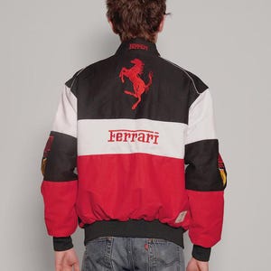 May include: A Ferrari racing jacket in black, white, and red. The jacket displays the Ferrari logo and text in red. The back of the jacket has a large Ferrari logo and the word "Ferrari" across the chest. It has black cuffs and a black collar.