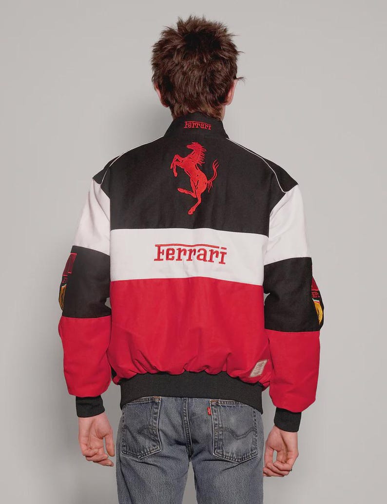 May include: A Ferrari racing jacket in black, white, and red. The jacket displays the Ferrari logo and text in red. The back of the jacket has a large Ferrari logo and the word "Ferrari" across the chest. It has black cuffs and a black collar.