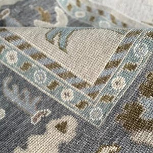 Könnte beinhalten: Nahaufnahme eines aufgerollten Teppichs mit detailliertem Muster. Der Teppich weist eine Kombination aus Grau-, Beige- und Blautönen auf. Das Design umfasst florale und geometrische Elemente mit einem Rand aus sich wiederholenden Mustern.