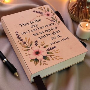 May include: A light pink journal with a black ribbon bookmark and a black pen. The cover features floral illustrations and the text: This is the day the Lord has made; let us rejoice and be glad in it. PSALM 118:24. The journal is rectangular.