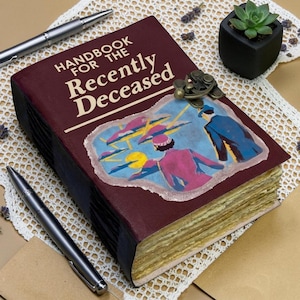 May include: A maroon hardcover book titled Handbook for the Recently Deceased with a vintage-style illustration of two figures on the cover. The book features a metal latch closure and thick, textured pages, accompanied by two silver pens.