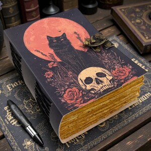 K&ouml;nnte beinhalten: Ein dekoratives Notizbuch mit schwarzem Einband, das eine schwarze Katzen-Silhouette vor einem roten Mond, einem Totenkopf und Rosen zeigt. Das Notizbuch hat alt aussehende Seiten und einen Metallverschluss. Ein Stift liegt auf einer passenden dekorativen Unterlage.