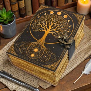 May include: A black journal with a gold tree of life design, moon phases, and a clasp. The journal is on a wooden surface with candles, a succulent plant, a pen, and a feather.