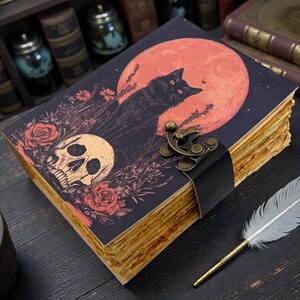 K&ouml;nnte beinhalten: Ein Tagebuch im Vintage-Stil mit einer Illustration einer schwarzen Katze vor einem roten Mond, einem Totenkopf und Rosen. Das Tagebuch hat gealtert aussehende Seiten, einen schwarzen Lederriemen und einen Metallverschluss. Eine Feder liegt daneben.