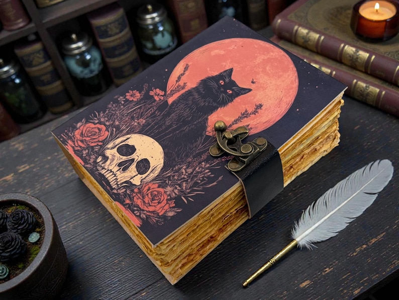 K&ouml;nnte beinhalten: Ein Tagebuch im Vintage-Stil mit einer Illustration einer schwarzen Katze vor einem roten Mond, einem Totenkopf und Rosen. Das Tagebuch hat gealtert aussehende Seiten, einen schwarzen Lederriemen und einen Metallverschluss. Eine Feder liegt daneben.