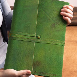 May include: A rectangular green leather journal with a textured surface and a wrap-around leather cord closure. The cover features the embossed text "My family art journal" in the bottom right corner, showcasing a natural finish and visible grain.