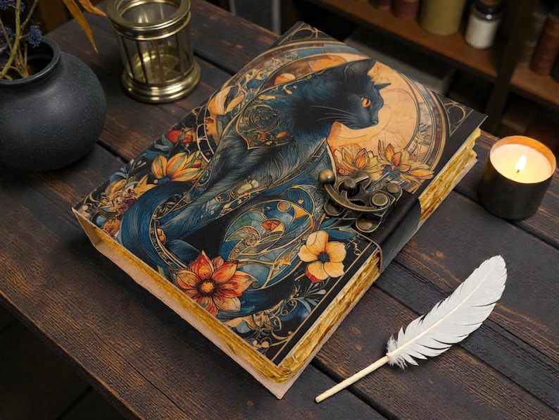 May include: A decorative journal with a black cat illustration on the cover, featuring orange and yellow floral accents. The journal has a clasp closure and aged-look pages. A white feather and lit candle are nearby.