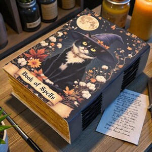May include: A decorative book titled "Book of Spells" featuring a black cat wearing a witch hat, surrounded by autumn leaves and a full moon. The book has aged-looking pages and is placed on a wooden surface.