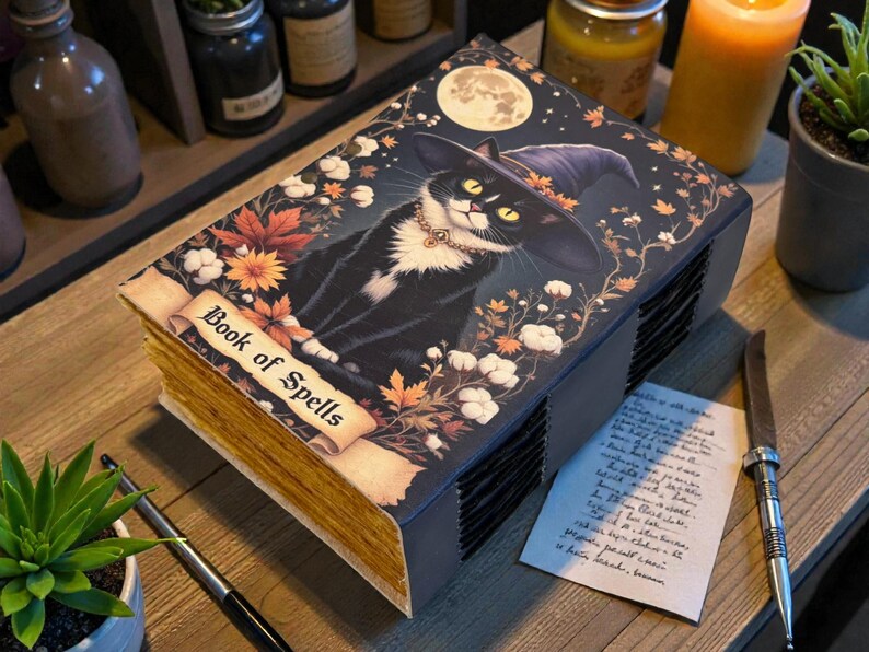 May include: A decorative book titled "Book of Spells" featuring a black cat wearing a witch hat, surrounded by autumn leaves and a full moon. The book has aged-looking pages and is placed on a wooden surface.