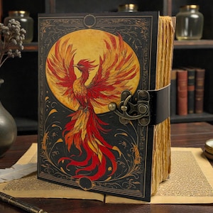 Peut inclure: Un journal de style vintage avec une illustration de ph&eacute;nix sur la couverture. La couverture est noire avec une bordure dor&eacute;e et une grande image d'un ph&eacute;nix rouge, orange et jaune sur un soleil jaune. Le journal est reli&eacute; par une sangle en cuir noir et un fermoir en m&eacute;tal.