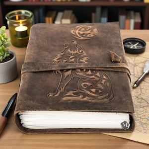 Peut inclure: Un journal en cuir marron avec un motif de loup en relief sur la couverture. Le journal est sécurisé par une lanière en cuir et présente un motif de lune. Le journal est ouvert, révélant des pages vierges.