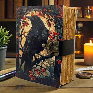 May include: A decorative journal with a raven illustration on the cover. The cover art features a black raven perched on a branch with red leaves, set against a stained-glass style background. The journal has a leather strap closure and aged paper.