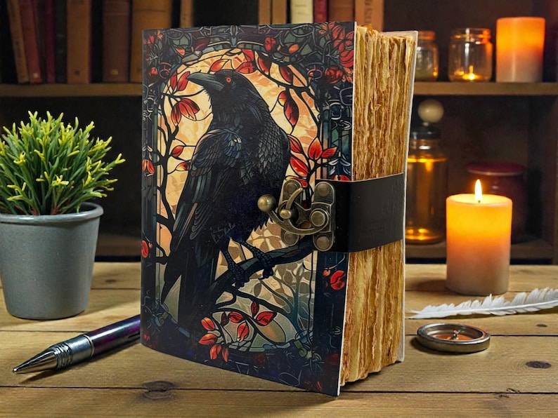May include: A decorative journal with a raven illustration on the cover. The cover art features a black raven perched on a branch with red leaves, set against a stained-glass style background. The journal has a leather strap closure and aged paper.