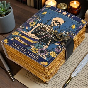 May include: A vintage-style journal with a blue cover featuring a skeleton reading a book, surrounded by flowers and moons. The journal has aged, yellowed pages and a clasp closure. The text "THE READER" is visible on the cover.