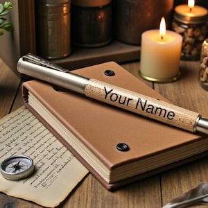 K&ouml;nnte beinhalten: Ein personalisierter Stift mit dem Text "Your Name" auf dem Holzschaft liegt auf einem braunen, ledergebundenen Notizbuch. Ein Kompass, ein handgeschriebener Brief, Kerzen und Gl&auml;ser befinden sich im Hintergrund und erzeugen eine Vintage-&Auml;sthetik.