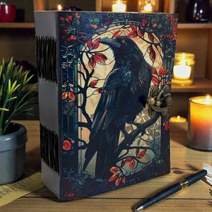 May include: A leather-bound journal with a raven illustration on the cover. The cover art features a black raven perched on a branch, surrounded by red and orange leaves, with a brass clasp. The journal is on a wooden surface.