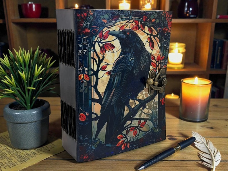 May include: A leather-bound journal with a raven illustration on the cover. The cover art features a black raven perched on a branch, surrounded by red and orange leaves, with a brass clasp. The journal is on a wooden surface.