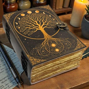 May include: A large, antique-style journal with a black cover featuring a gold tree of life design, moon phases, and stars. The pages have a golden hue. A pen and handwritten notes are on the wooden surface.