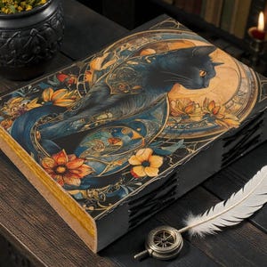 May include: A decorative book with a black cat illustration on the cover, featuring orange and yellow flowers and a blue background. A white feather and a compass-like object are next to the book. The book has a black spine and gold-colored pages.