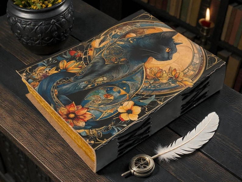 May include: A decorative book with a black cat illustration on the cover, featuring orange and yellow flowers and a blue background. A white feather and a compass-like object are next to the book. The book has a black spine and gold-colored pages.
