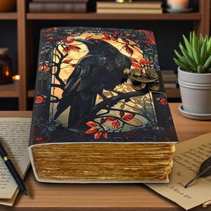 May include: A decorative journal with a raven illustration on the cover. The cover features a black raven perched on a branch with red leaves, set against a yellow background. The journal has a metal clasp and aged-looking pages.