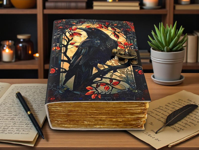 May include: A decorative journal with a raven illustration on the cover. The cover features a black raven perched on a branch with red leaves, set against a yellow background. The journal has a metal clasp and aged-looking pages.