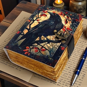 May include: A decorative journal with a raven illustration on the cover. The cover features a black raven perched on a branch with red and orange leaves. The journal has a leather strap closure and aged-looking pages. A blue pen rests on the paper next to the journal.