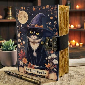 May include: A decorative journal with a black cat wearing a witch hat on the cover, the words "Book of Spells" are printed on a banner. The journal has a black leather strap and clasp. The pages have a golden hue.
