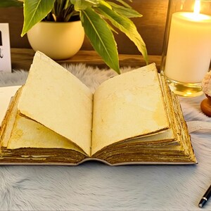 K&ouml;nnte beinhalten: Ein offenes, gealtertes Buch mit vergilbten Seiten liegt auf einer flauschigen, grauen Oberfl&auml;che. Eine brennende Kerze in einem Glas, eine kleine Pflanze und ein Stift befinden sich in der N&auml;he. Die Seiten des Buches sind fleckig.