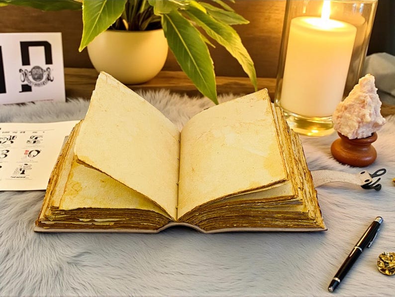 K&ouml;nnte beinhalten: Ein offenes, gealtertes Buch mit vergilbten Seiten liegt auf einer flauschigen, grauen Oberfl&auml;che. Eine brennende Kerze in einem Glas, eine kleine Pflanze und ein Stift befinden sich in der N&auml;he. Die Seiten des Buches sind fleckig.