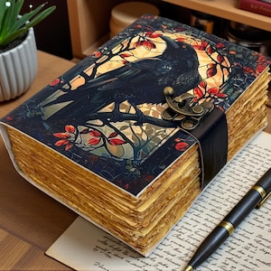 May include: A decorative journal with a raven illustration on the cover. The cover features a black raven perched on a branch, set against a stained-glass window design with red and gold accents. The journal has aged-looking pages and a clasp.