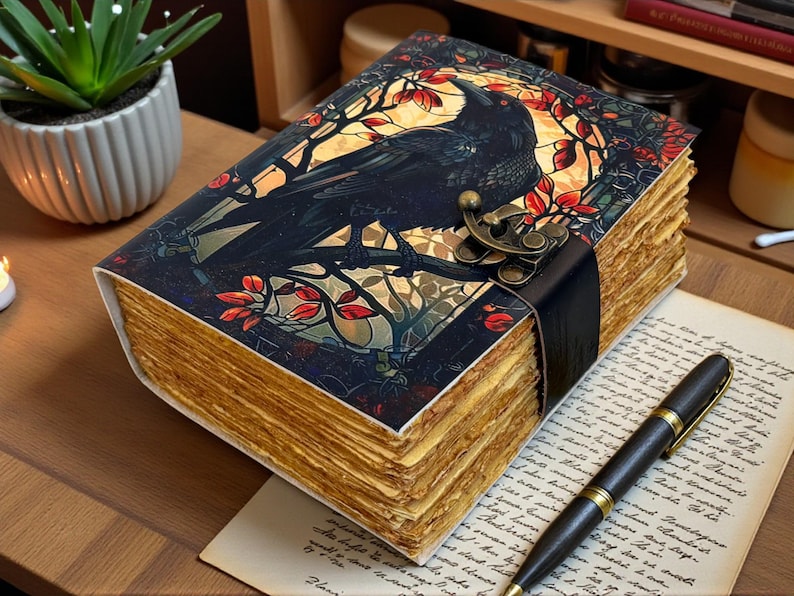 May include: A decorative journal with a raven illustration on the cover. The cover features a black raven perched on a branch, set against a stained-glass window design with red and gold accents. The journal has aged-looking pages and a clasp.