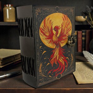 Peut inclure: Un journal en cuir noir avec une illustration de ph&eacute;nix. La couverture pr&eacute;sente un ph&eacute;nix rouge, orange et jaune vif sur un fond de lune jaune. Le journal a un fermoir en m&eacute;tal et des coutures d&eacute;coratives le long du dos. Un stylo et une plume sont sur la surface en bois.