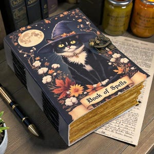 May include: A decorative book titled "Book of Spells" featuring a black cat wearing a witch hat, surrounded by autumn leaves and a full moon. The book has aged-looking pages and a clasp.