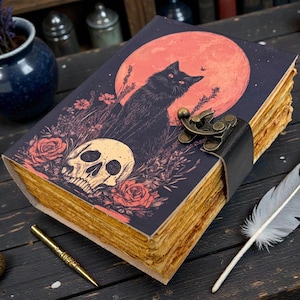 K&ouml;nnte beinhalten: Ein dekoratives Notizbuch mit einer Illustration einer schwarzen Katze auf dem Cover, mit einem Totenkopf und Rosen. Das Notizbuch hat einen Lederriemenverschluss und alt aussehende Seiten. Eine Feder und ein Stift liegen neben dem Notizbuch.