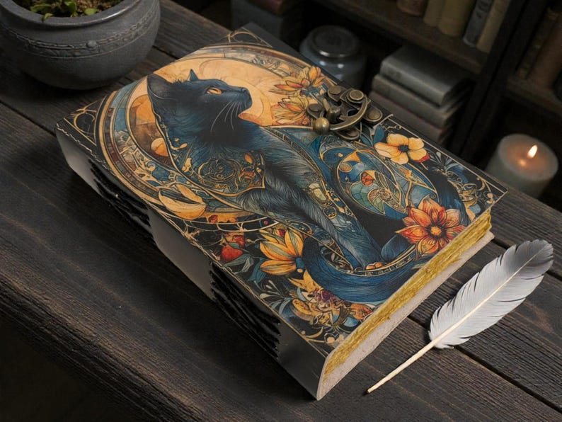 May include: A decorative journal with a black cat illustration on the cover, surrounded by floral and celestial designs. The journal has a metal clasp and gold-edged pages. A white feather quill rests beside it on a dark wooden surface.
