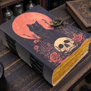 K&ouml;nnte beinhalten: Ein dekoratives Buch mit einer Illustration einer schwarzen Katze vor einem roten Mond, einem Totenkopf und floralen Elementen. Das Buch hat goldfarbene Kanten und einen Metallverschluss. Das Thema ist gotisch und mystisch.