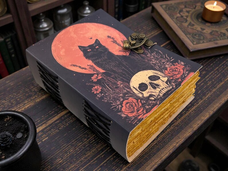 K&ouml;nnte beinhalten: Ein dekoratives Buch mit einer Illustration einer schwarzen Katze vor einem roten Mond, einem Totenkopf und floralen Elementen. Das Buch hat goldfarbene Kanten und einen Metallverschluss. Das Thema ist gotisch und mystisch.
