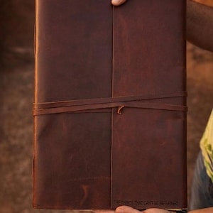 May include: A dark brown leather journal with a smooth, matte finish and a wrap-around leather tie closure. The bottom right corner of the front cover features the embossed text, "THE THINGS THAT CAN'T BE RETURNED," in a simple, clean font.