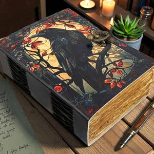 May include: A decorative journal with a raven illustration on the cover. The cover features a stained-glass style design with a black raven perched on a branch. The pages have a gold-colored edge. A pen rests on the wooden surface.