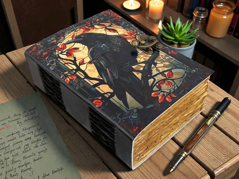 May include: A decorative journal with a raven illustration on the cover. The cover features a stained-glass style design with a black raven perched on a branch. The pages have a gold-colored edge. A pen rests on the wooden surface.